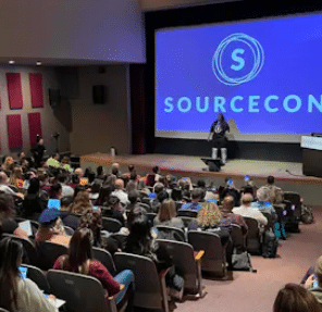 A speaker presents on stage at the SourceCon conference event to an audience of attentive attendees